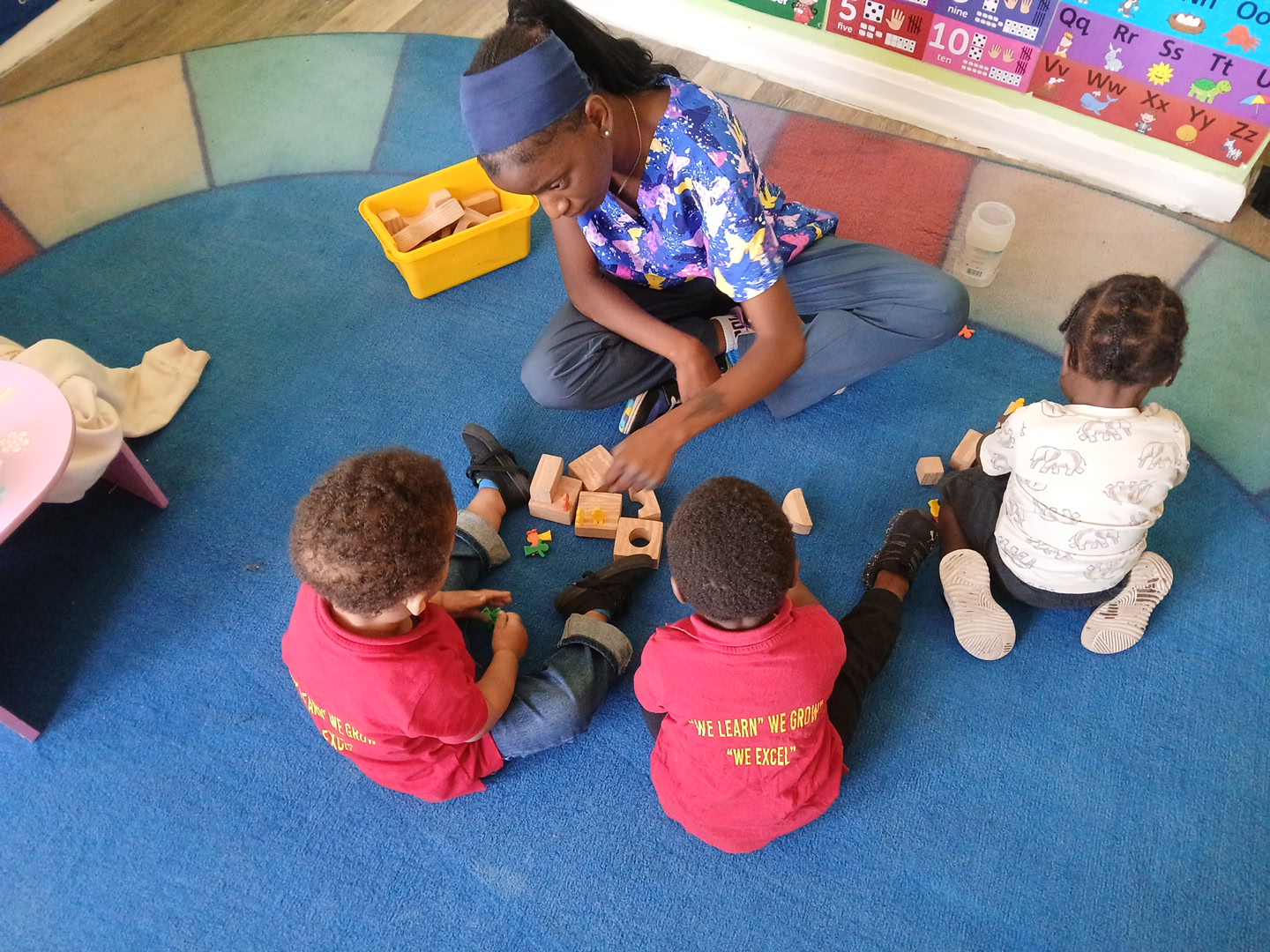 A group of four children standing in front of a white wall, with a large blank picture frame above them and potted plant b...
