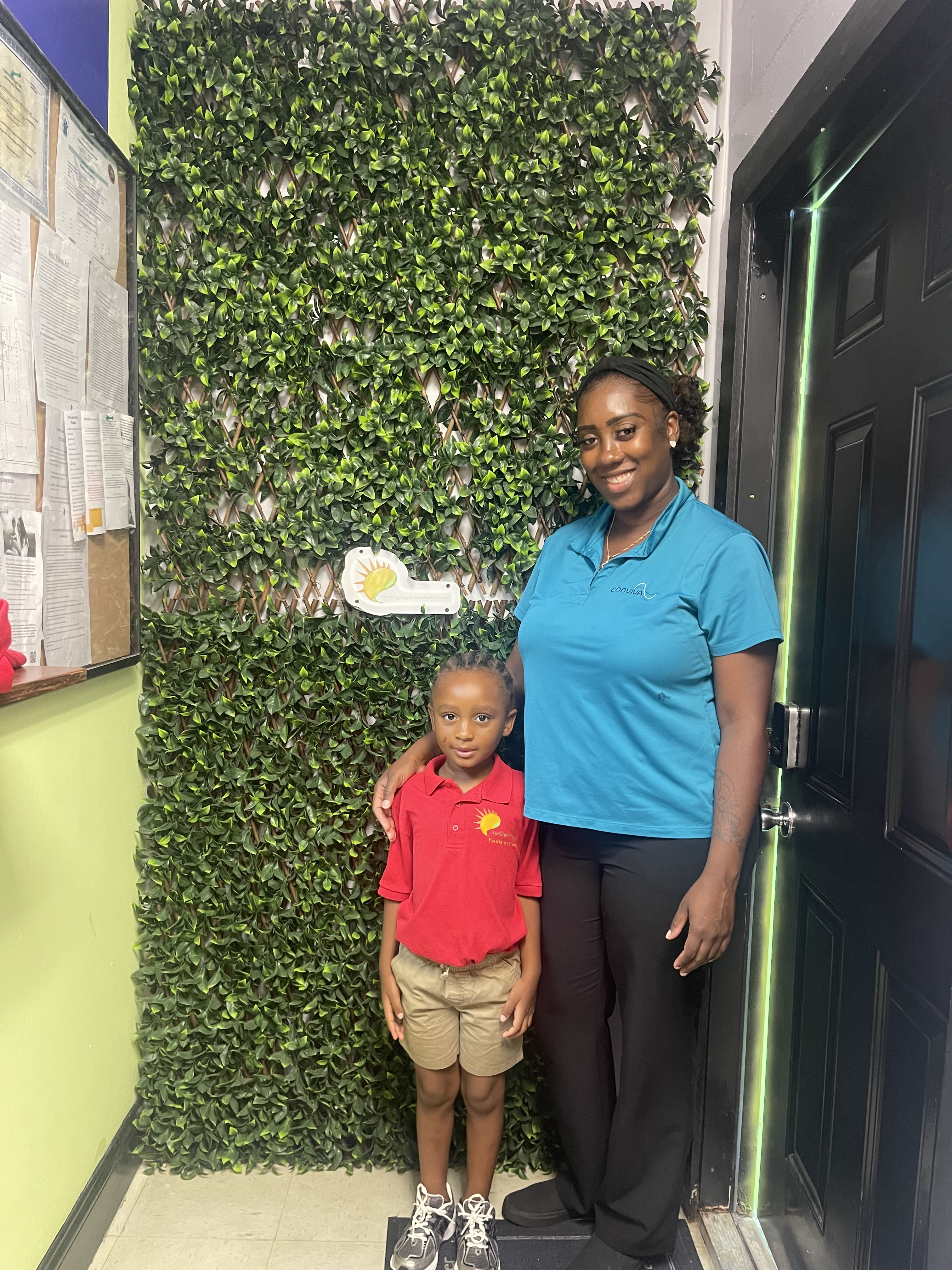 A group of four children standing in front of a white wall, with a large blank picture frame above them and potted plant b...